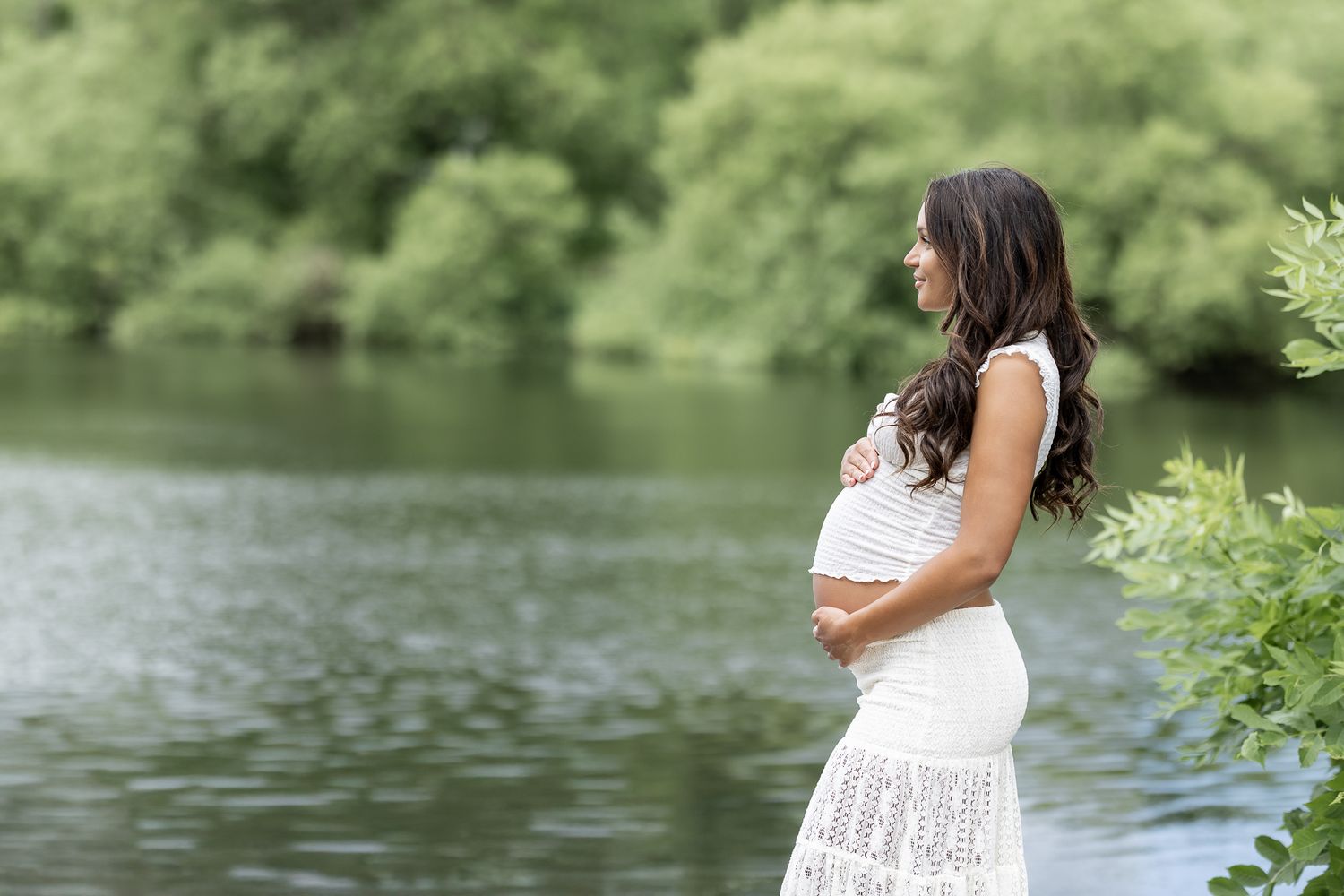 Pregnant woman in white dress cradling her baby bump beside a serene lake surrounded by lush green trees.