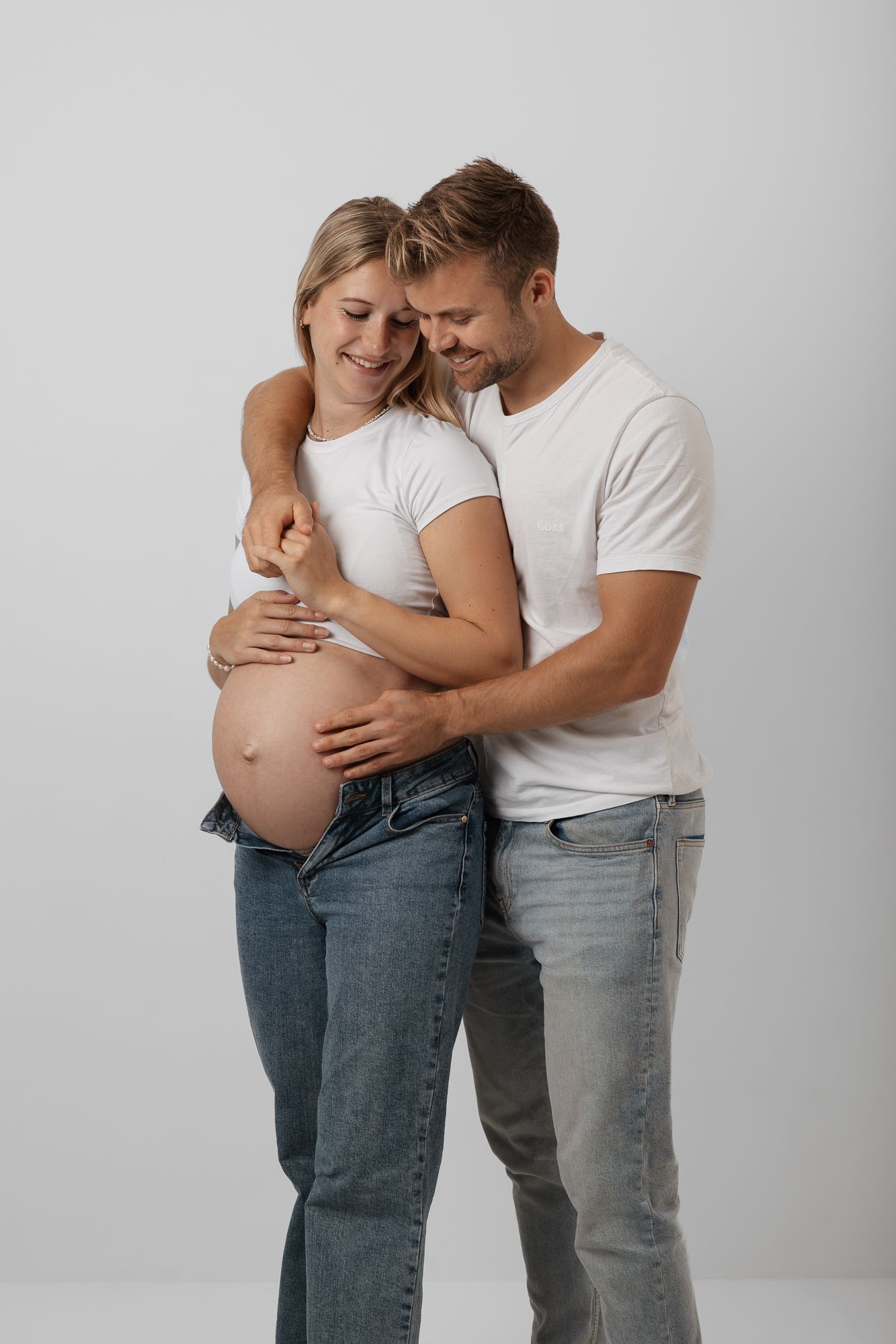Expectant couple in white shirts and jeans embrace lovingly while showing off pregnancy bump against white backdrop.