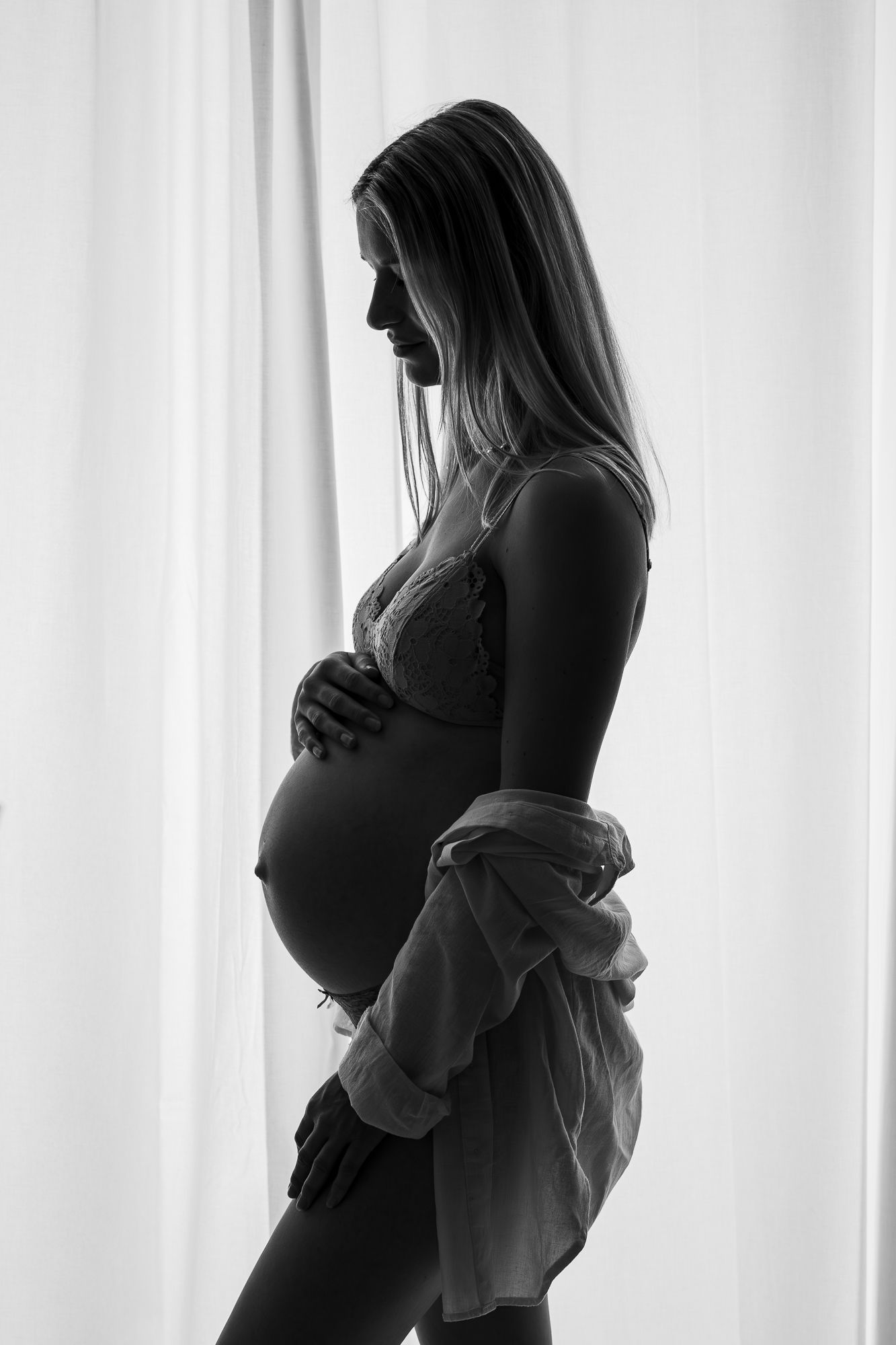 Silhouette of a pregnant woman in a flowing robe standing by a window with soft natural light.