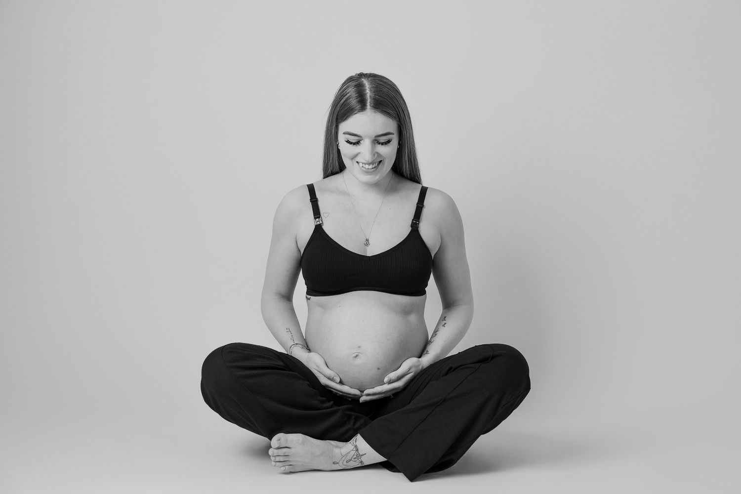A pregnant woman in black relaxation wear sits cross-legged on the floor in a minimalist black and white photo.