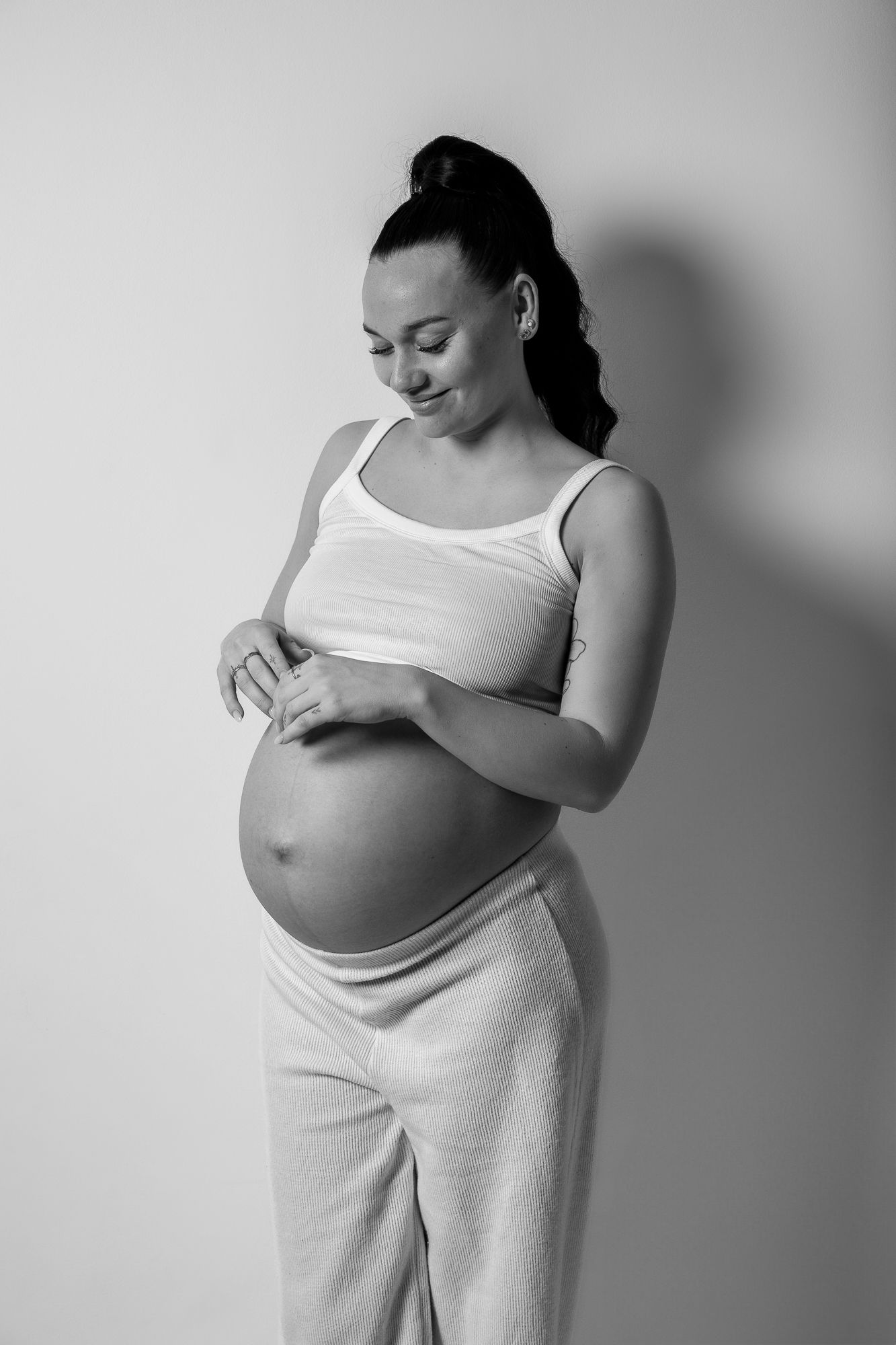 A black and white maternity portrait showing an expectant mother in white attire tenderly touching her pregnant belly.