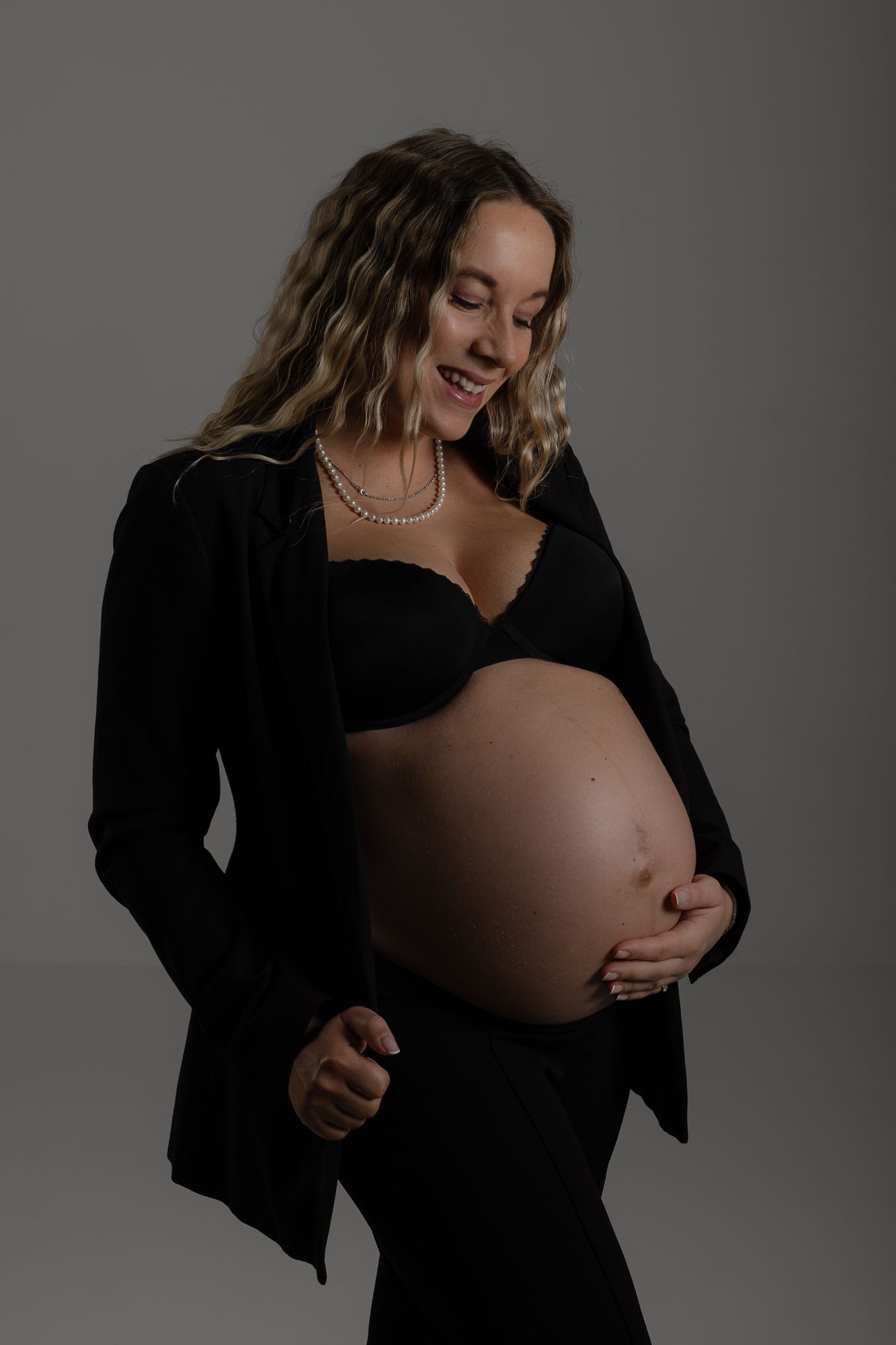 A maternity portrait of an expectant mother in black clothing against a dark backdrop, touching her pregnant belly.