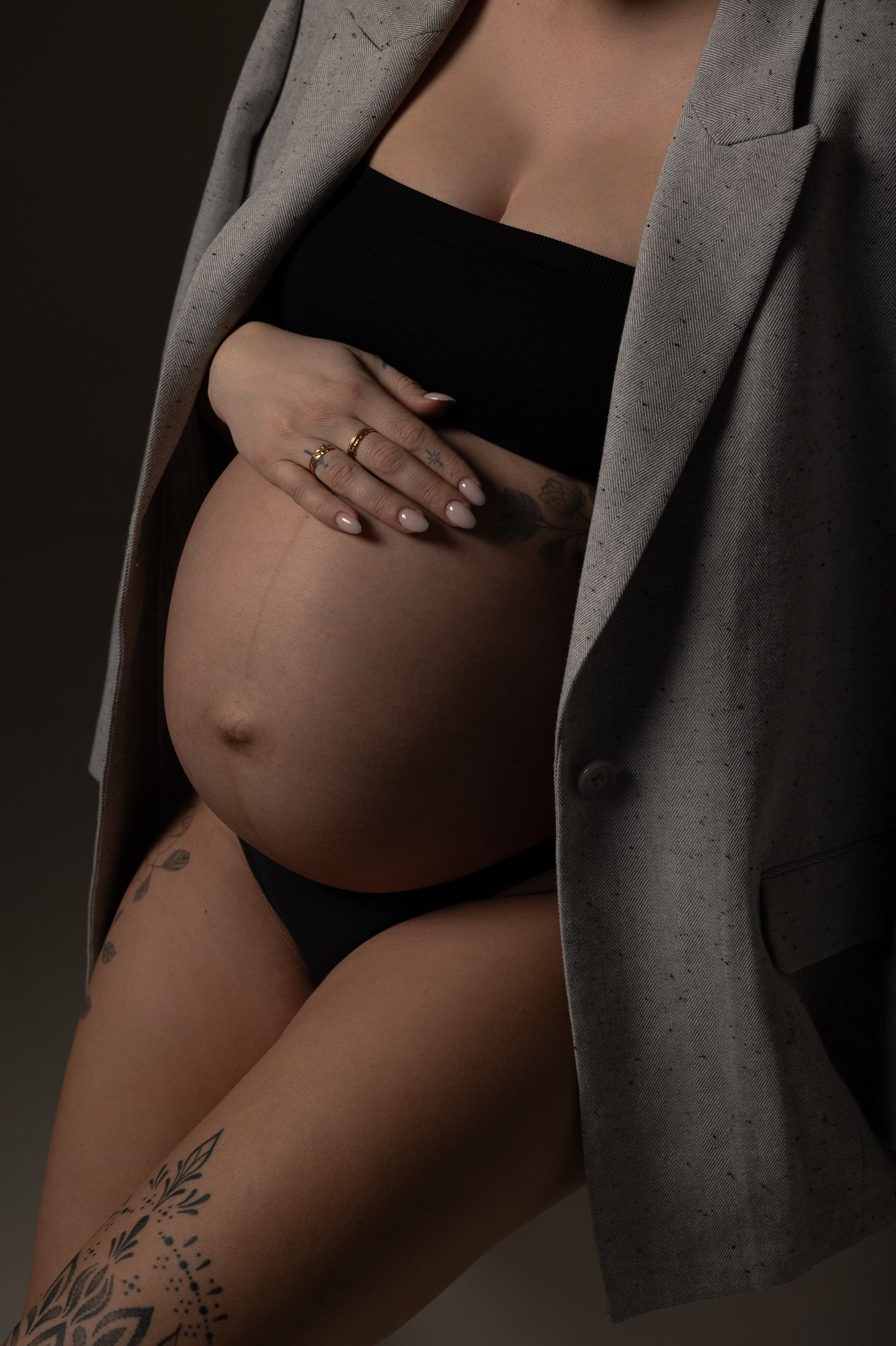 Artistic maternity photo showing pregnant belly under gray blazer with hand resting on bump and visible tattoo on thigh.