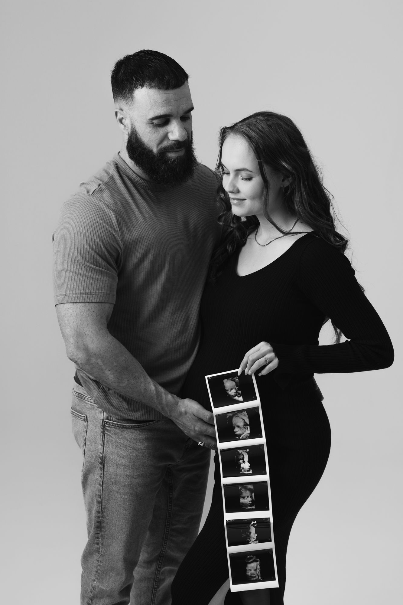 Black and white maternity photo showing a couple holding ultrasound photos in a vertical strip against a pregnant belly.