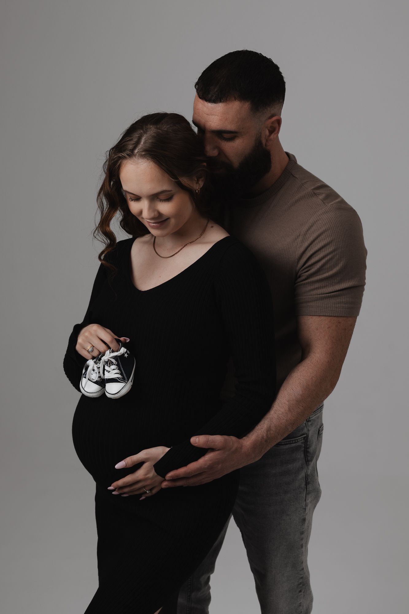 Expecting couple holds tiny baby shoes while embracing in intimate maternity photoshoot against gray backdrop.