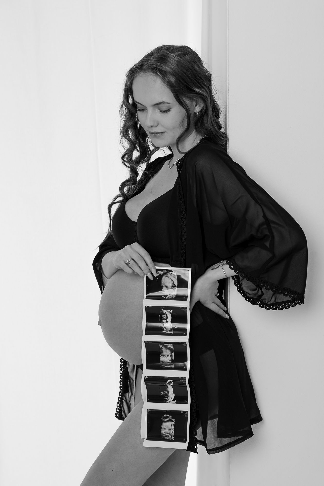 Black and white maternity photo showing pregnant woman in dark robe holding strip of ultrasound images.