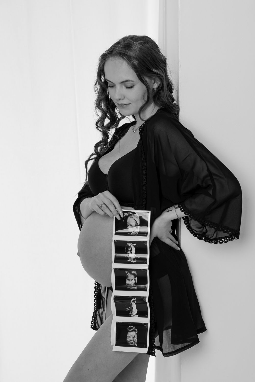 Black and white maternity photo showing pregnant woman in dark robe holding strip of ultrasound images.