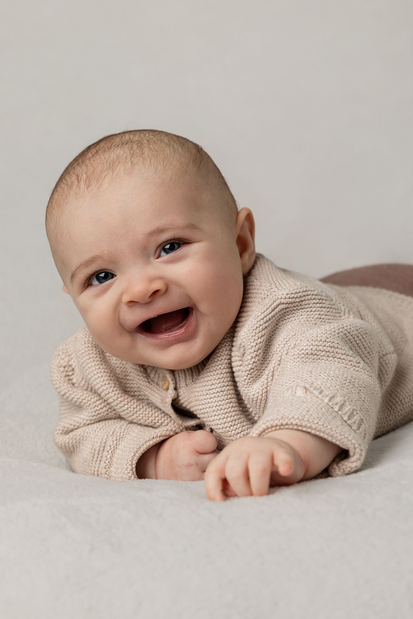 A sweet baby wearing a beige knit sweater smiles and lies on a neutral gray background in a series of photos.