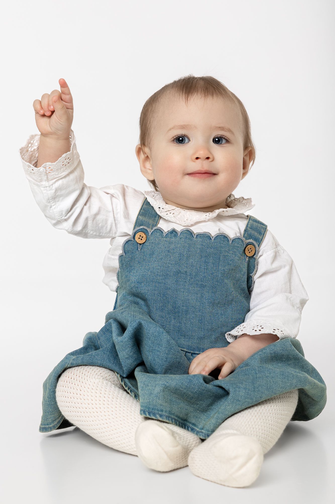 A small child wearing a white blouse and blue denim overalls sits on white backdrop while pointing upward.