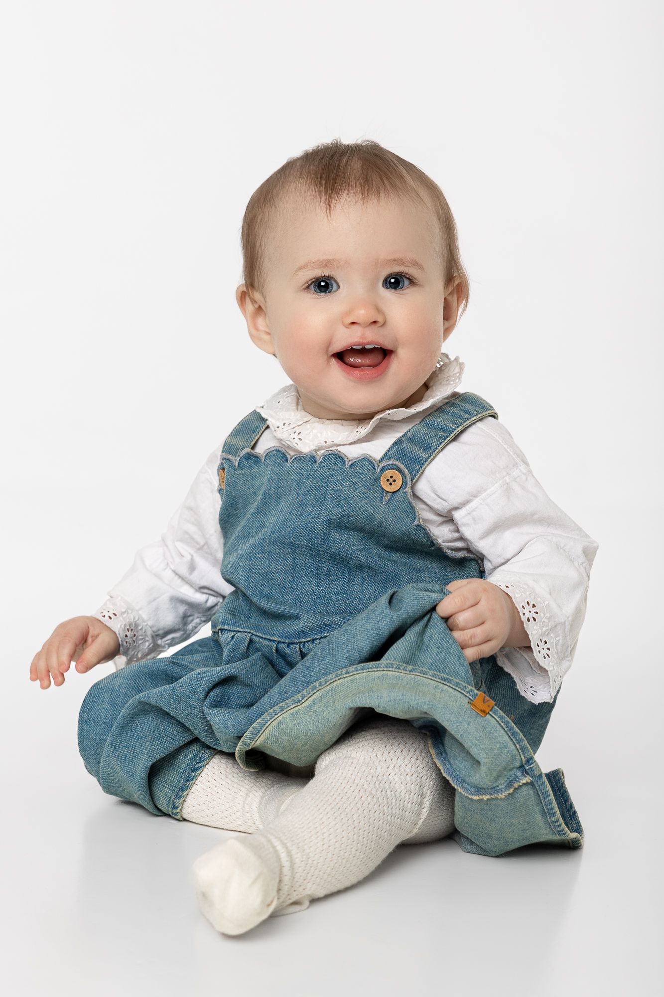 Little one wearing denim overalls with white shirt and white socks sits joyfully on white background.