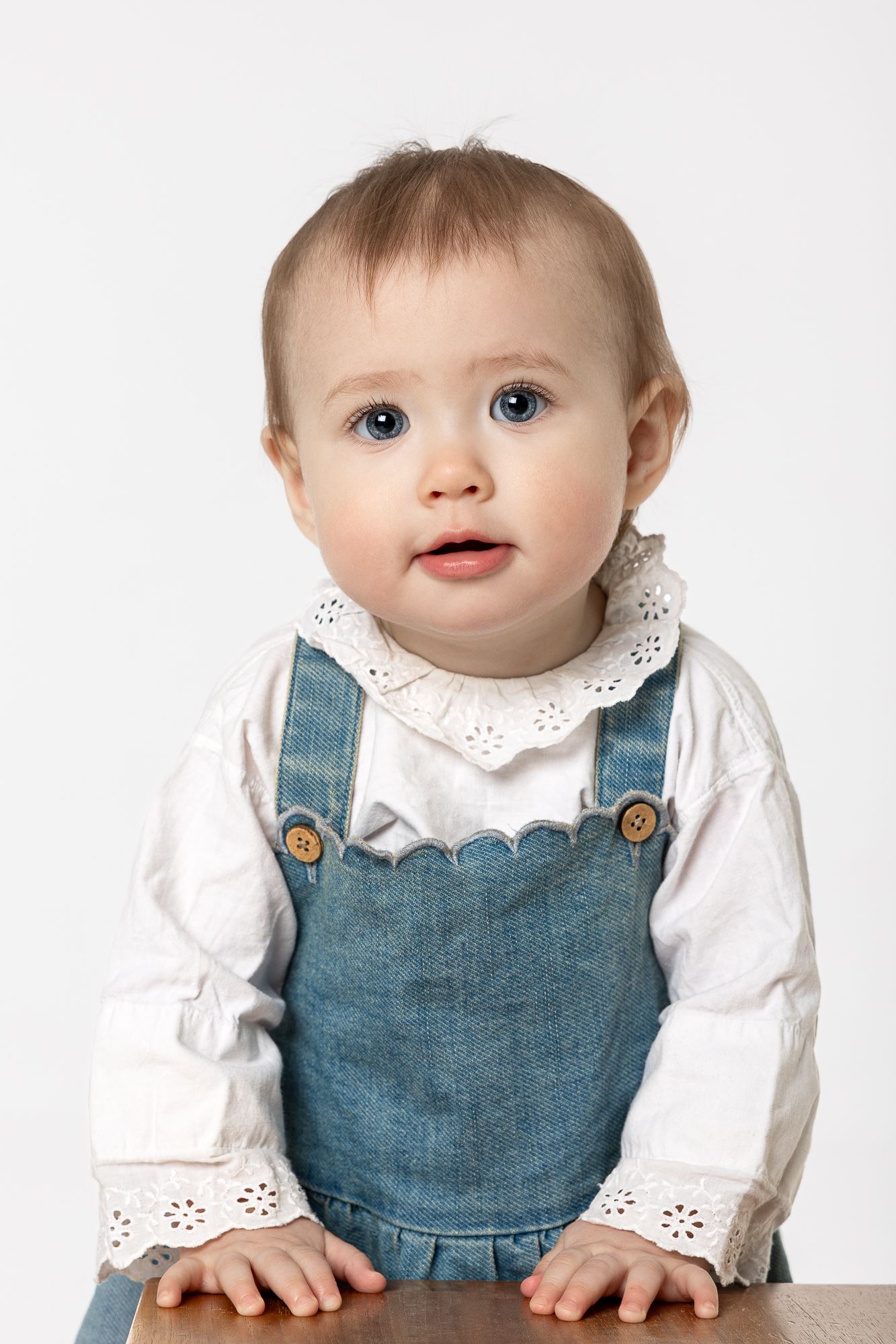 Baby wearing white eyelet blouse and denim overalls poses against white background.