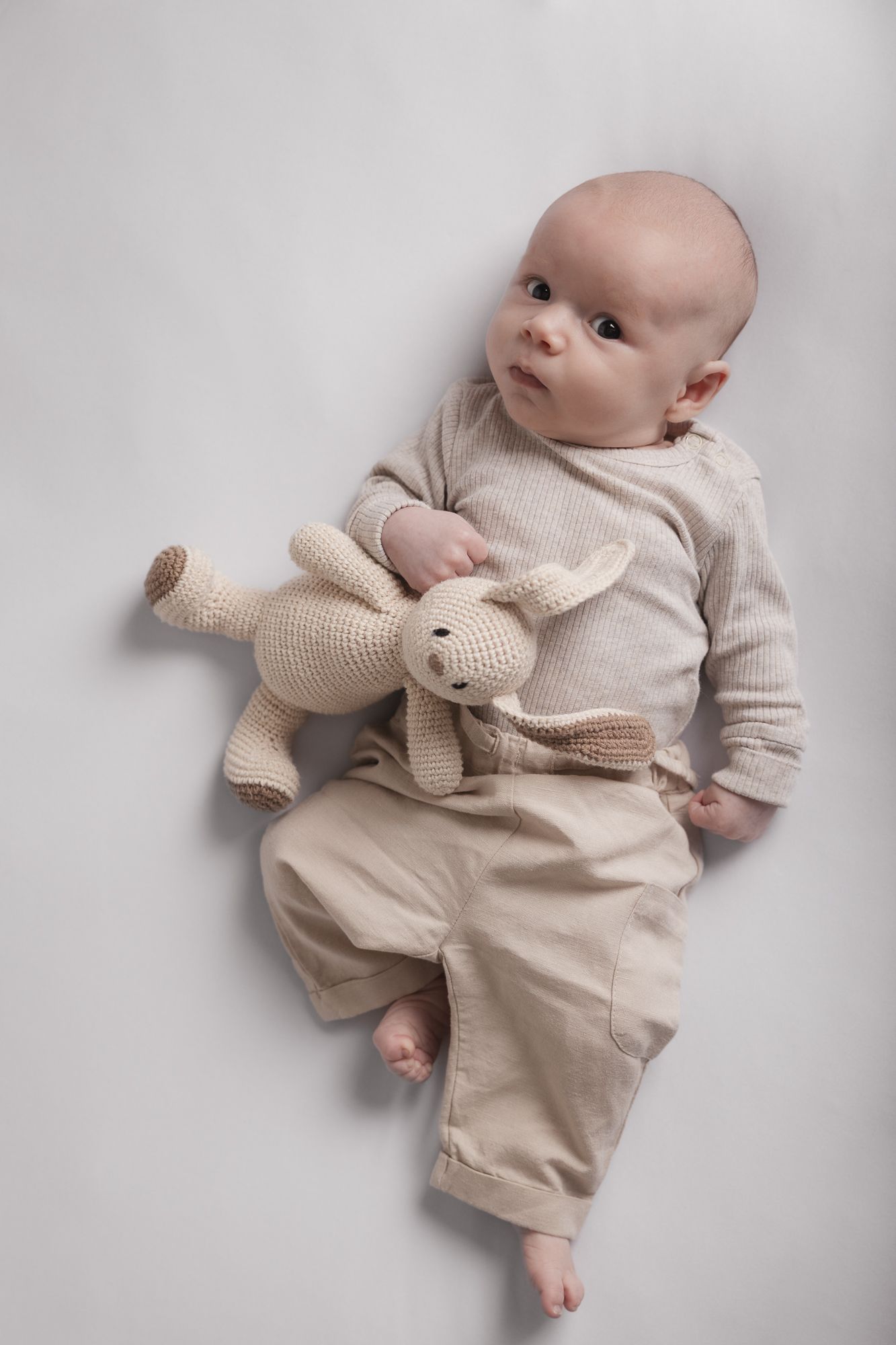 Small baby in neutral beige outfit lying on white background with matching knit stuffed animal toy.