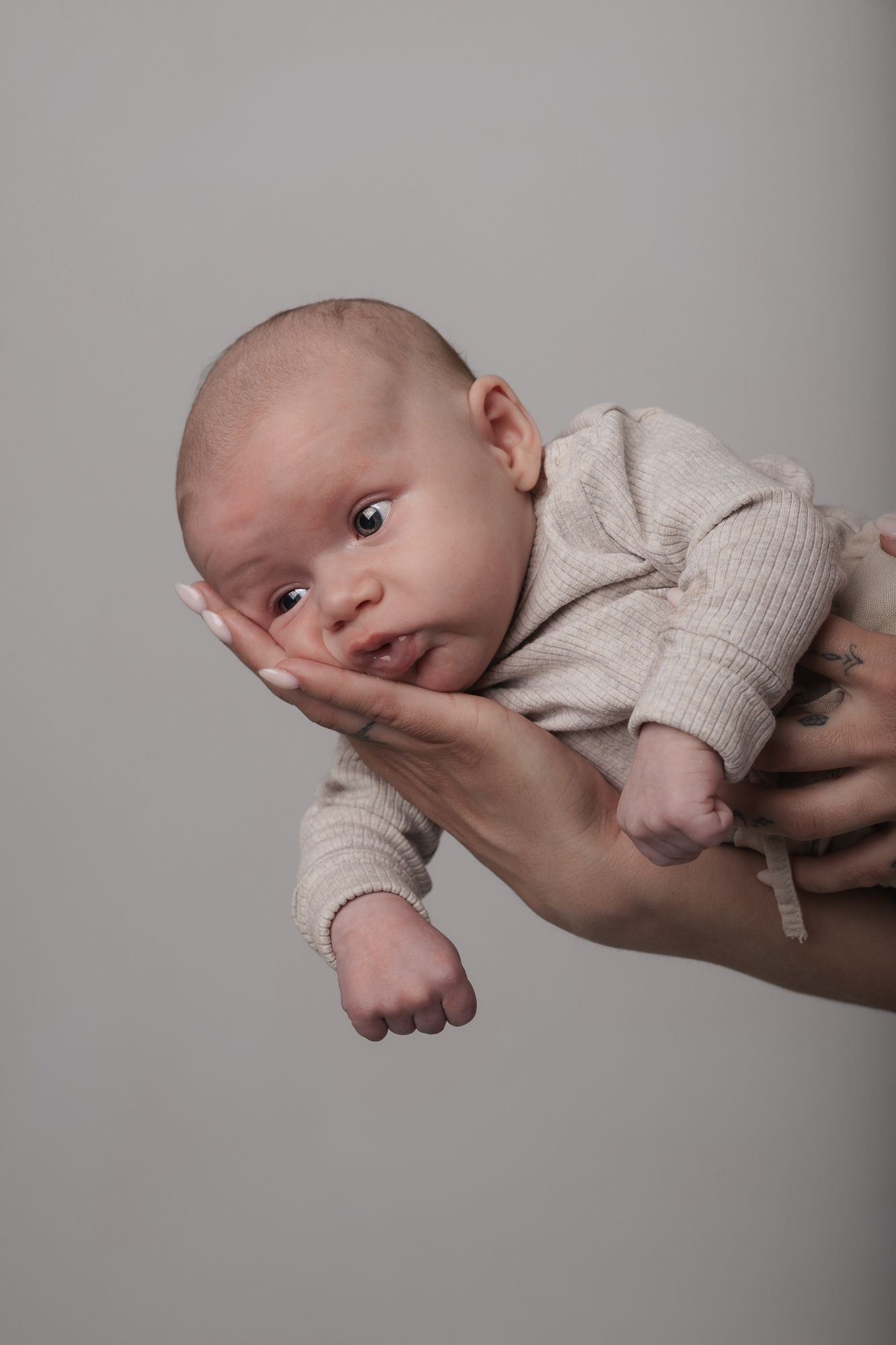 A small infant wearing a cream-colored outfit is gently supported by adult hands against a gray background.