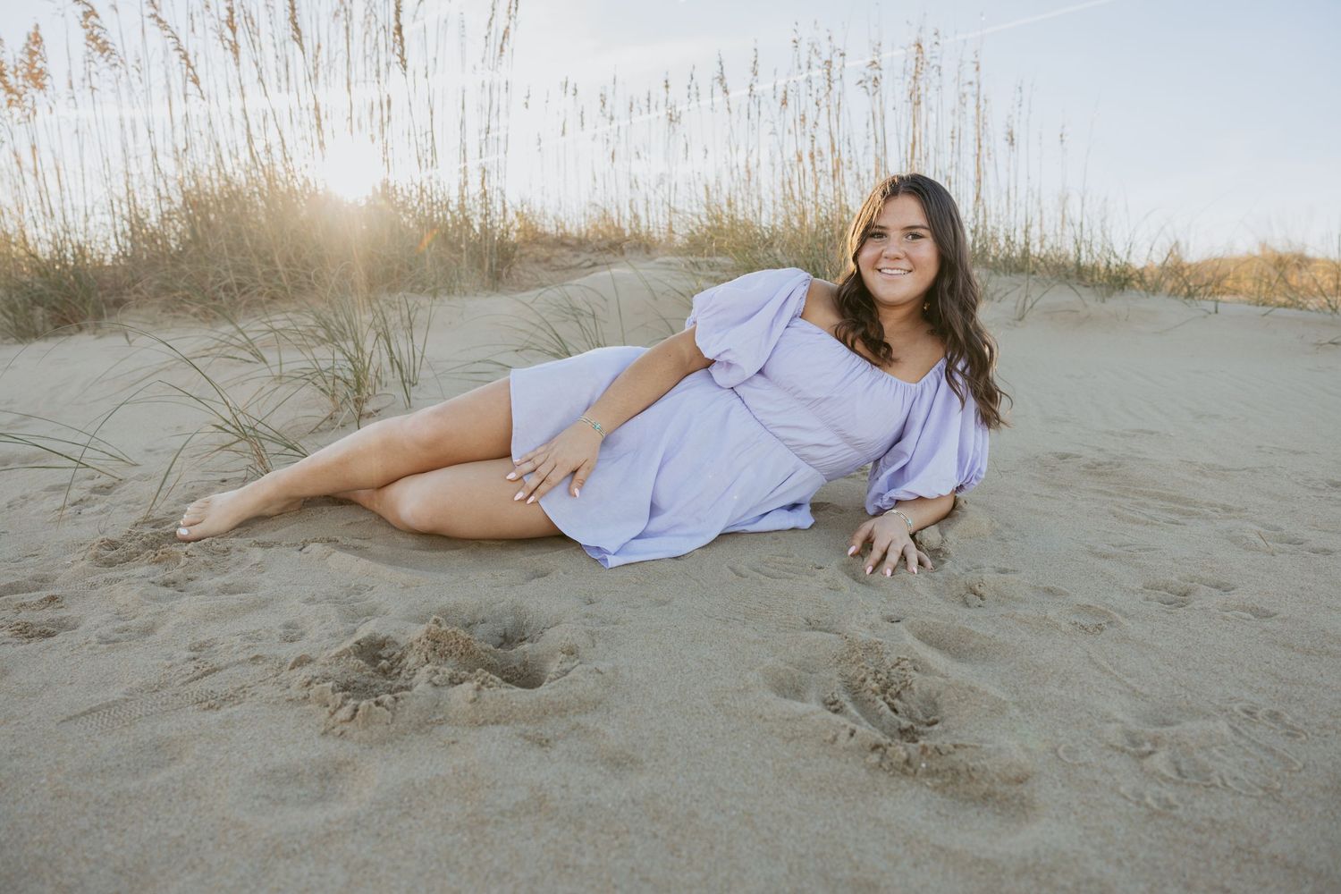 Someone in a light purple dress poses on a sandy beach with tall grass in the background during golden hour.