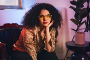 Confident woman with curly hair and glasses sitting on a couch under colorful lighting.