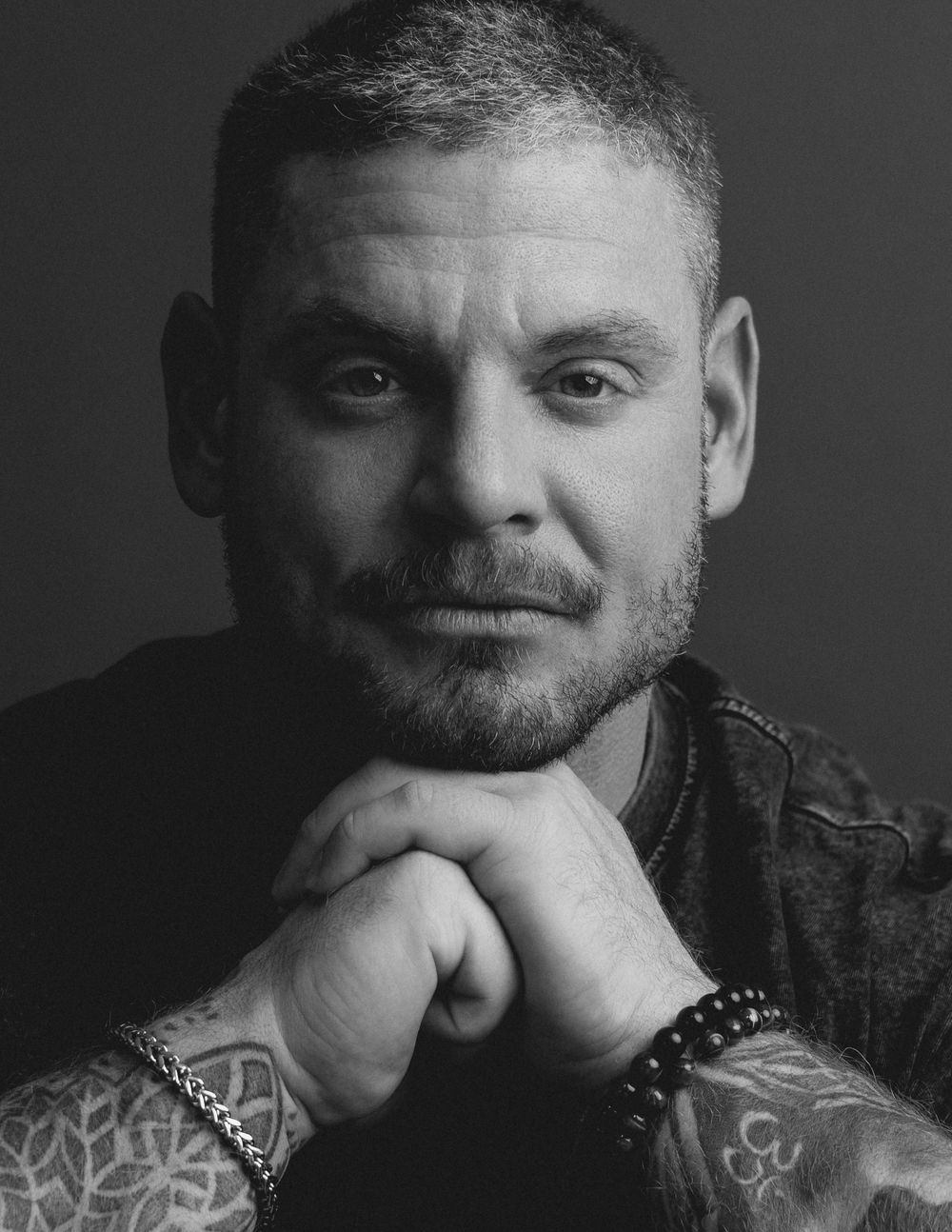Black-and-white portrait of a man with tattoos resting his chin on his hands.