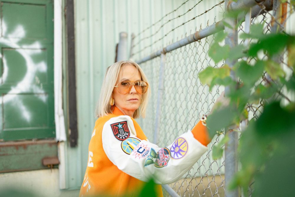 Stylish woman in orange sweater and sunglasses leaning on a chain-link fence outdoors.