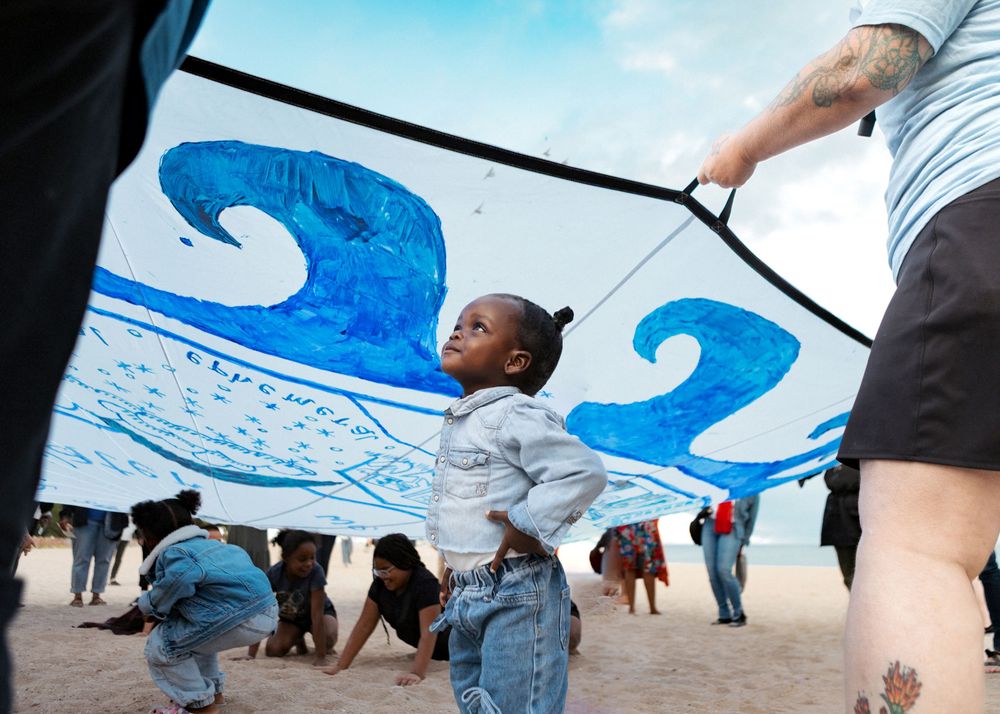 Child stands under large blue wave banner on sandy beach during outdoor community event.