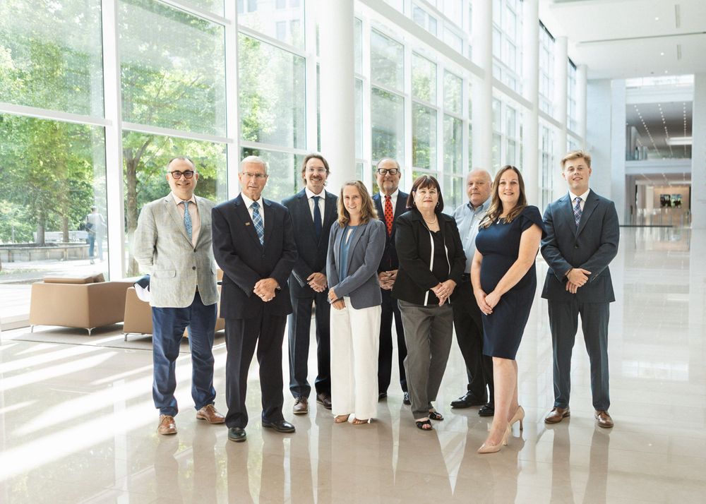 Group of nine business professionals posing together in a modern corporate lobby with floor-to-ceiling windows.