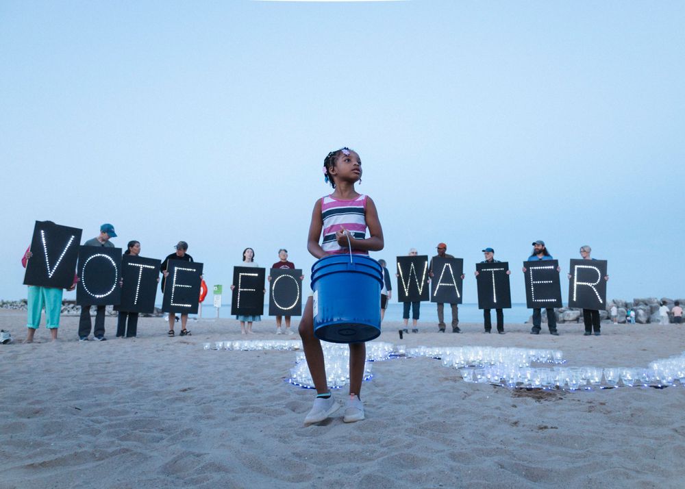 Figure stands on beach at dusk with protest signs visible in background.