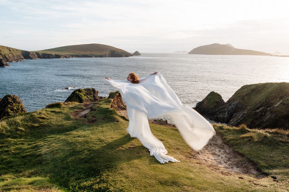 A flowing white wedding veil catches the wind against a dramatic coastal landscape at sunset.