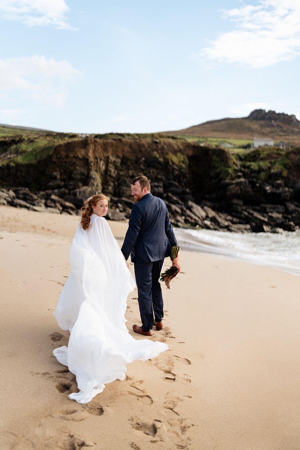 A couple walks along a scenic beach with rocky cliffs, the bride's white dress flowing in the coastal breeze.