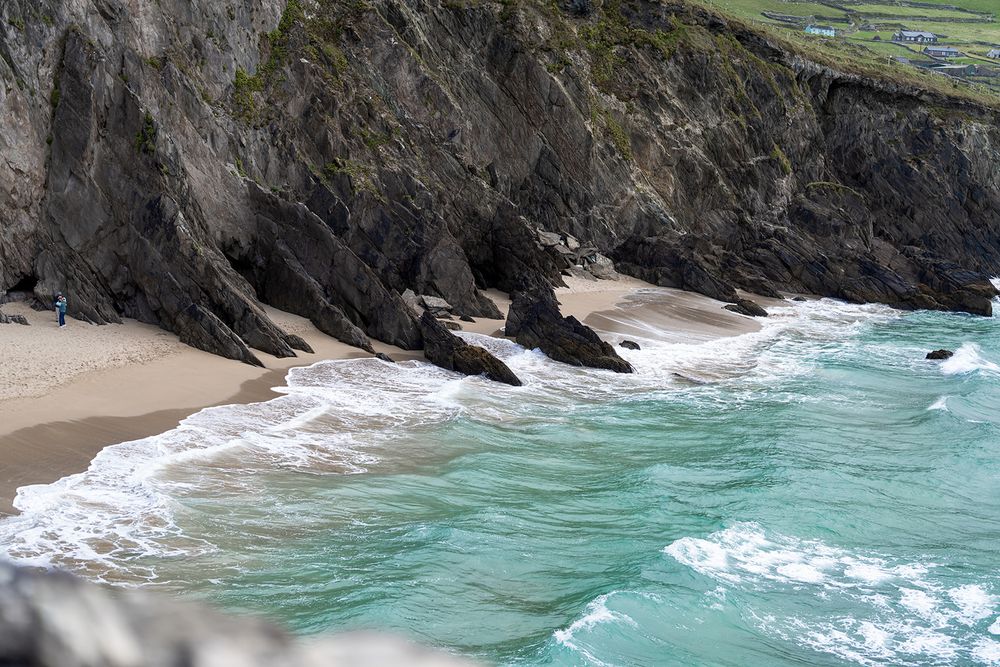 Turquoise ocean waves crash against rugged rocky cliffs along a dramatic coastal shoreline during a sunny day.