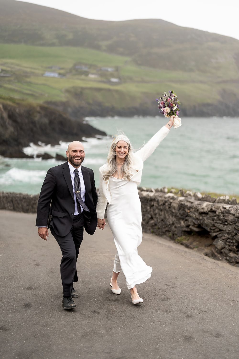 A couple walks joyfully along a coastal path in Ireland with dramatic cliffs and ocean views in the background.
