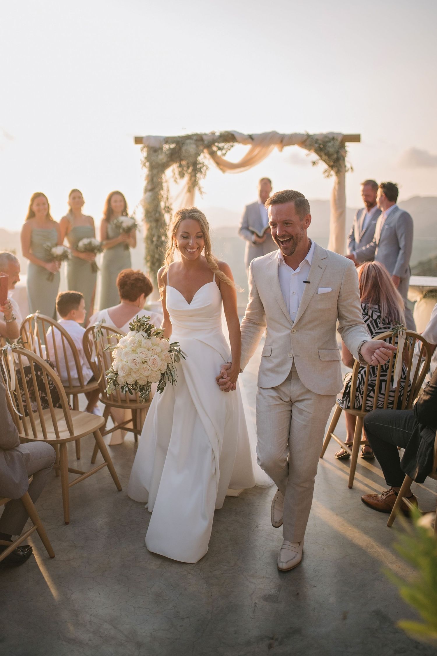 Couple walks down aisle at outdoor beach wedding ceremony with rustic wooden chairs and floral arch.