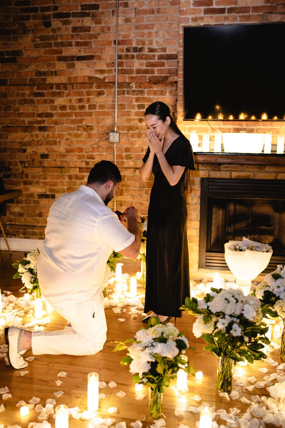 Man proposes on one knee surrounded by candles and white flowers as woman covers her mouth in surprise.
