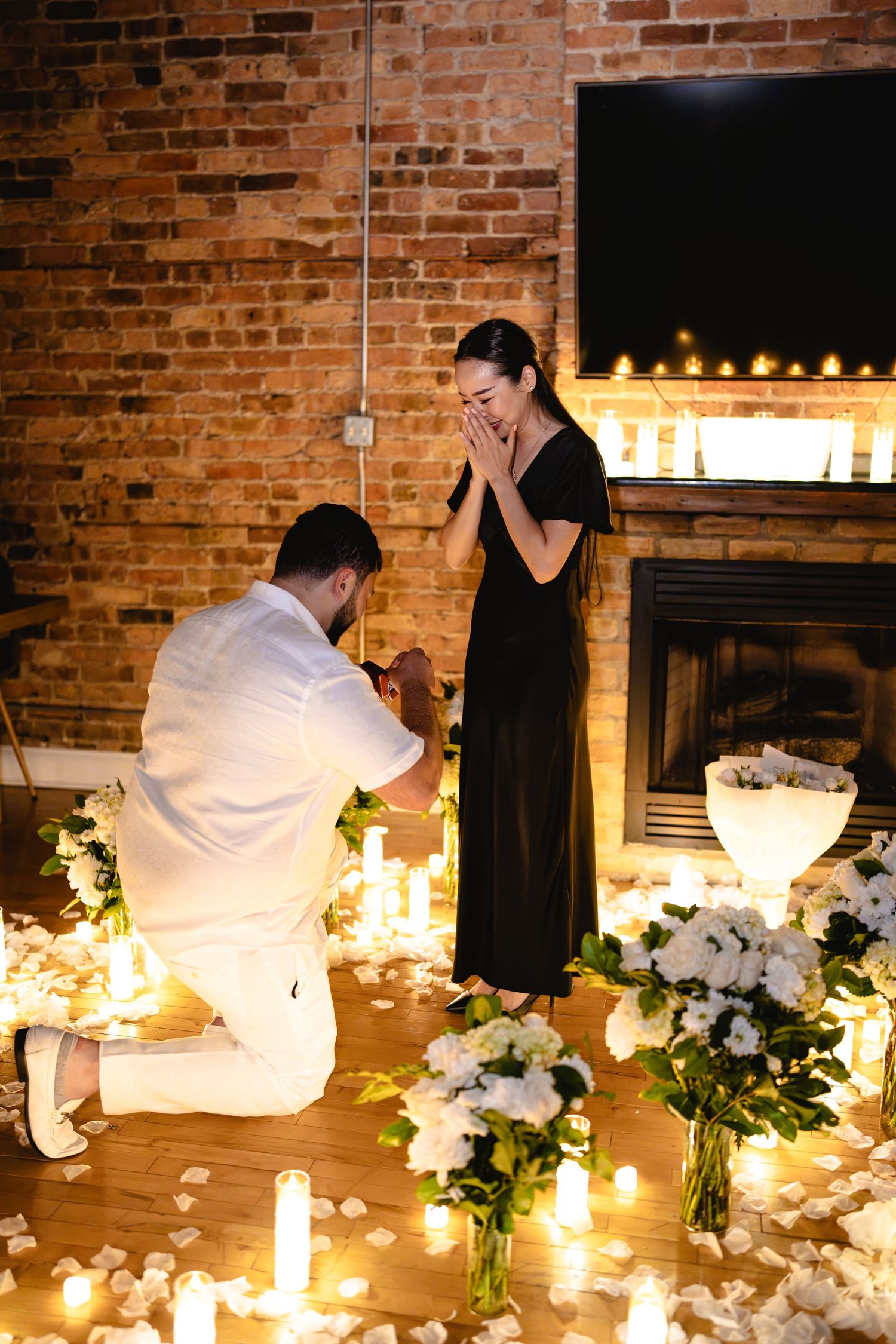 Man proposes on one knee surrounded by candles and white flowers as woman covers her mouth in surprise.