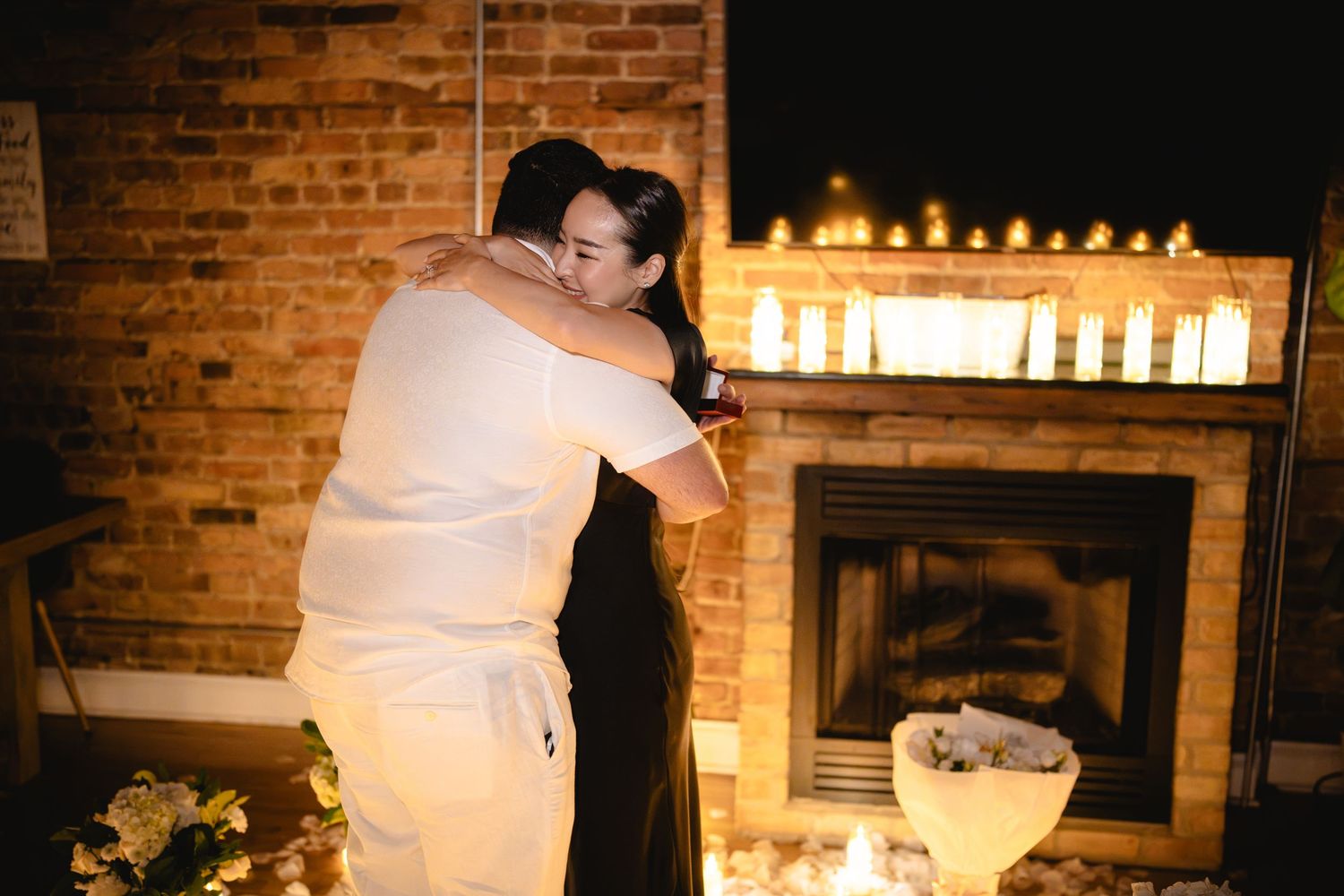 Couple embracing romantically by a candlelit brick fireplace in a dimly lit room at night.