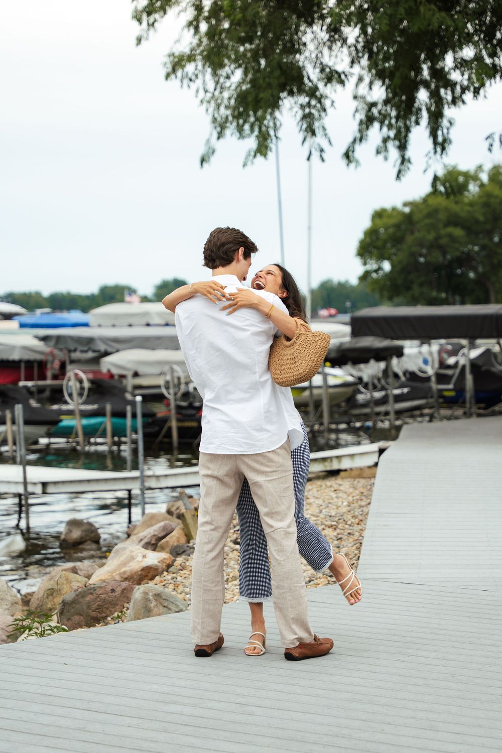 Couple embracing and spinning joyfully on a wooden dock near a marina with boats and water in the background.