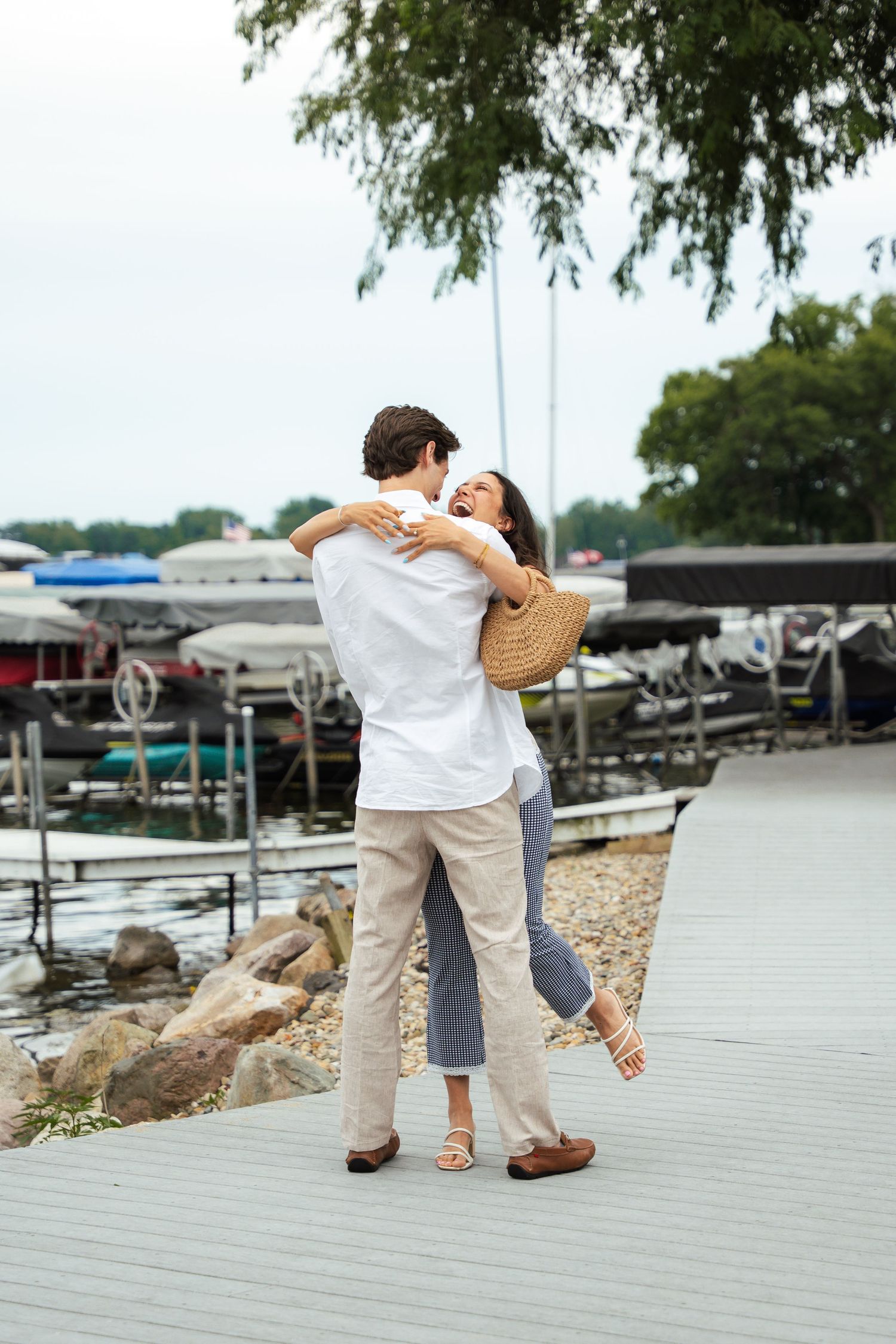 Couple embracing and spinning joyfully on a wooden dock near a marina with boats and water in the background.