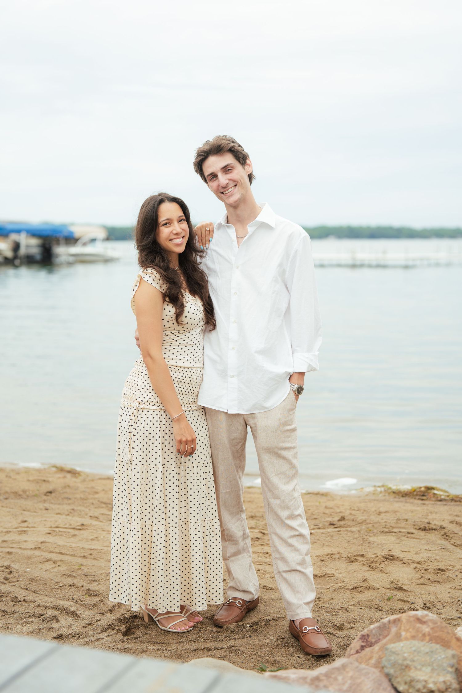Smiling couple posing on sandy beach, woman in polka dot dress and man in white shirt, with lake and marina in background.