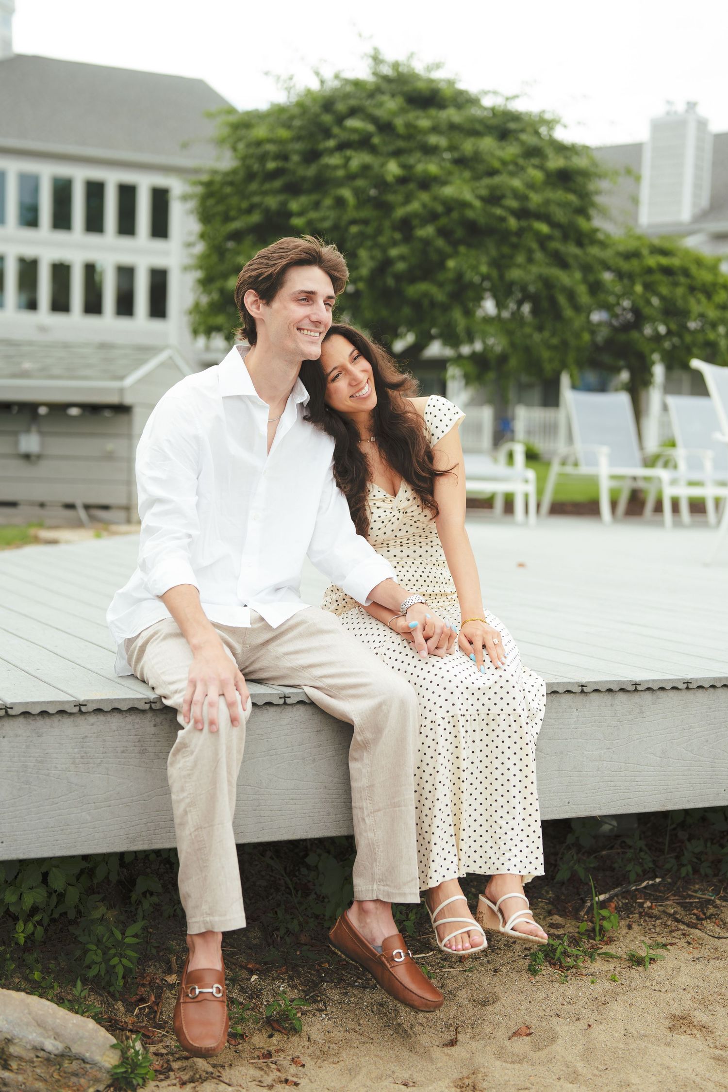 A smiling couple sits on a dock, man in white shirt and khakis, woman in polka dot dress, with a house and trees behind.