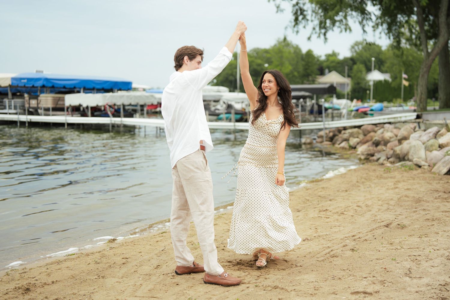 Couple dancing on sandy beach by waterfront, woman in flowing dress, man in white shirt, holding hands raised.