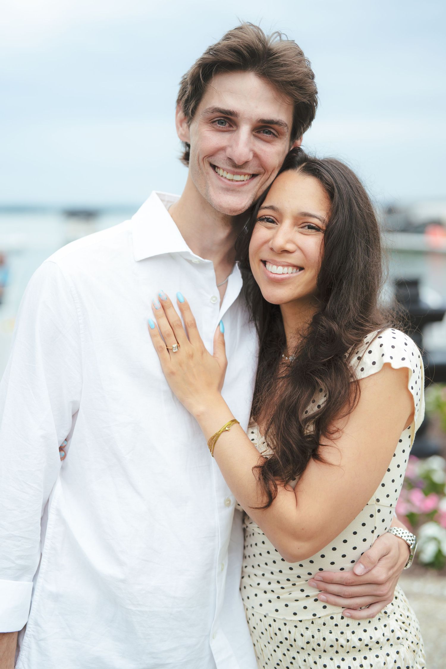 Happy couple embracing outdoors near waterfront, woman in polka dot dress showing engagement ring, man in white shirt.