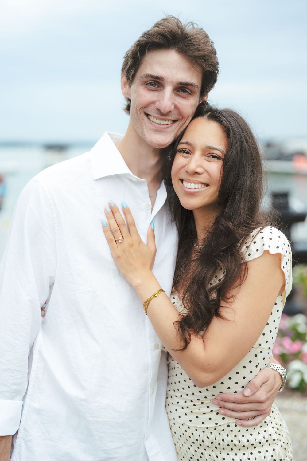 Happy couple embracing outdoors near waterfront, woman in polka dot dress showing engagement ring, man in white shirt.