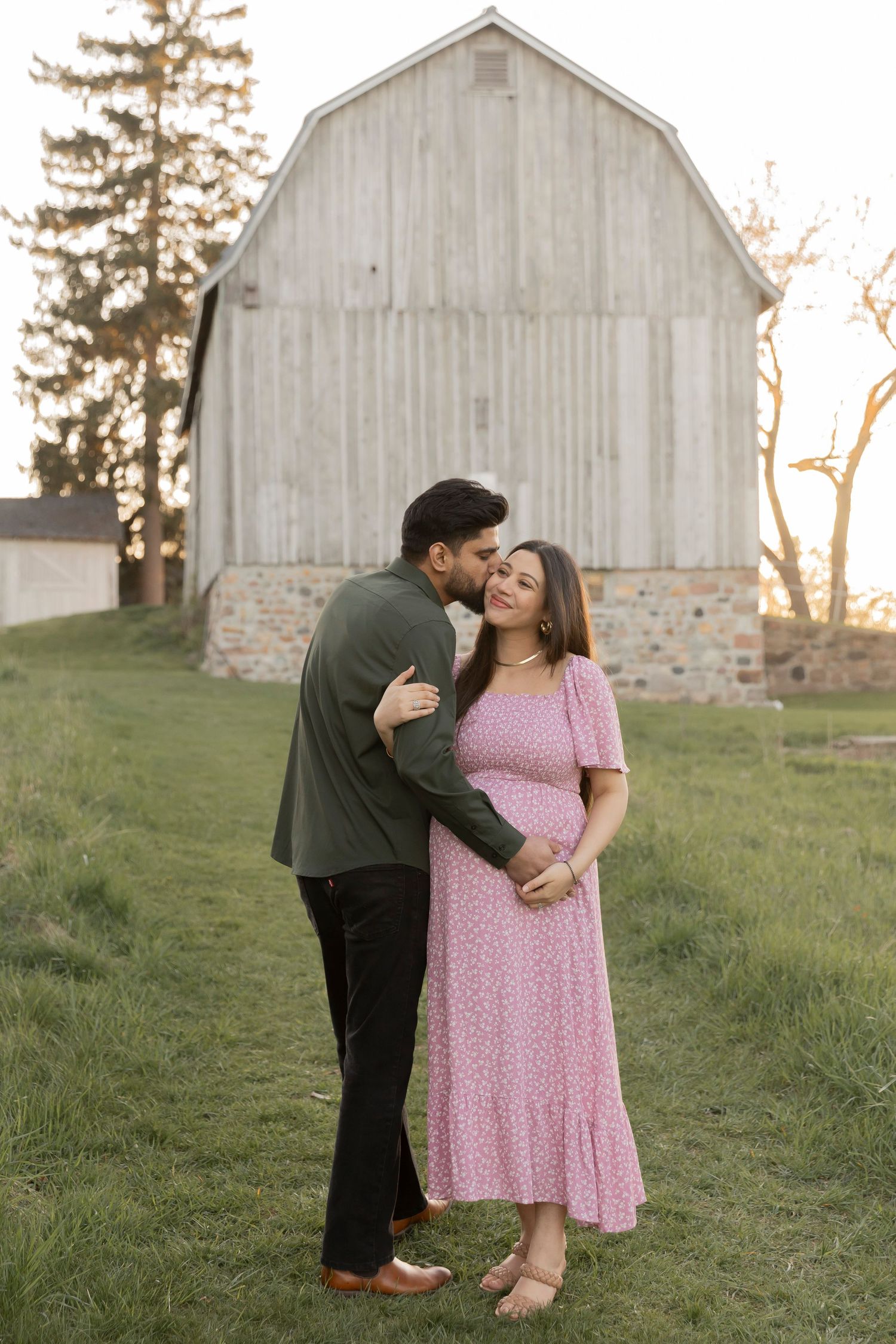 pregnant couple holding the belly and kissing in front of a barn in Oakland County michigan