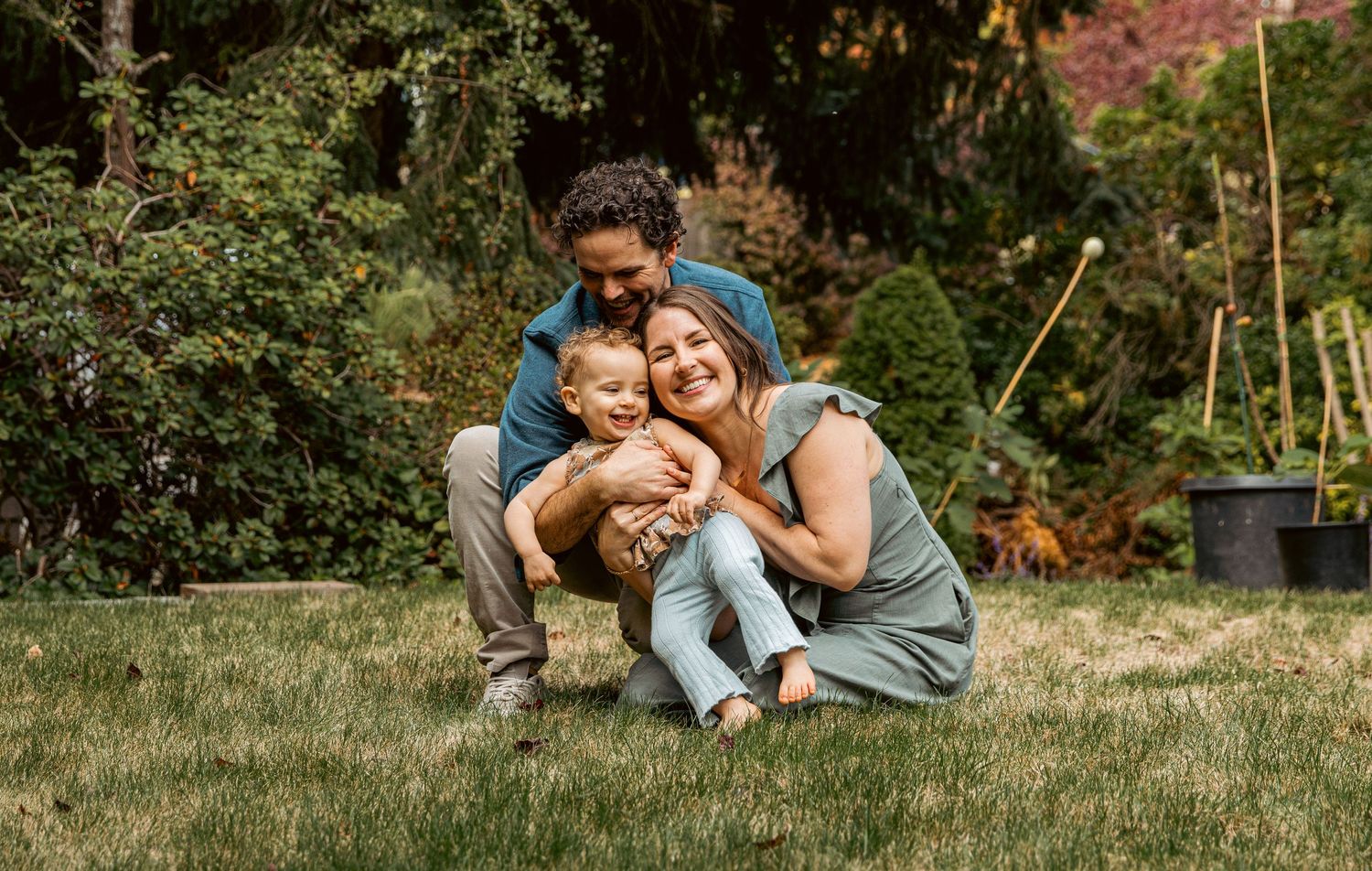 A happy family of three cuddles together on a green lawn surrounded by garden trees.