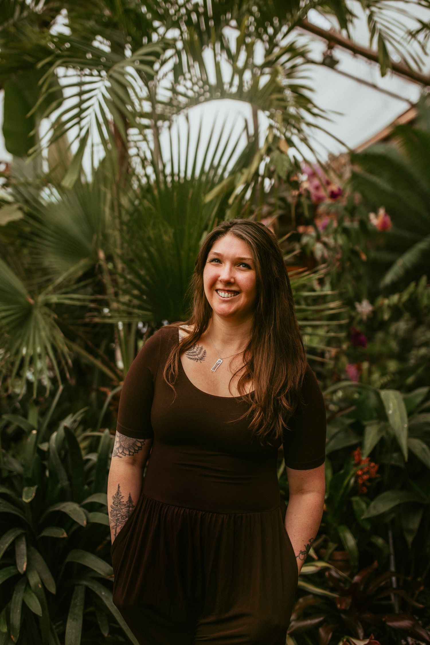 Smiling woman with tattoos in brown dress posing among tropical plants in a greenhouse setting.