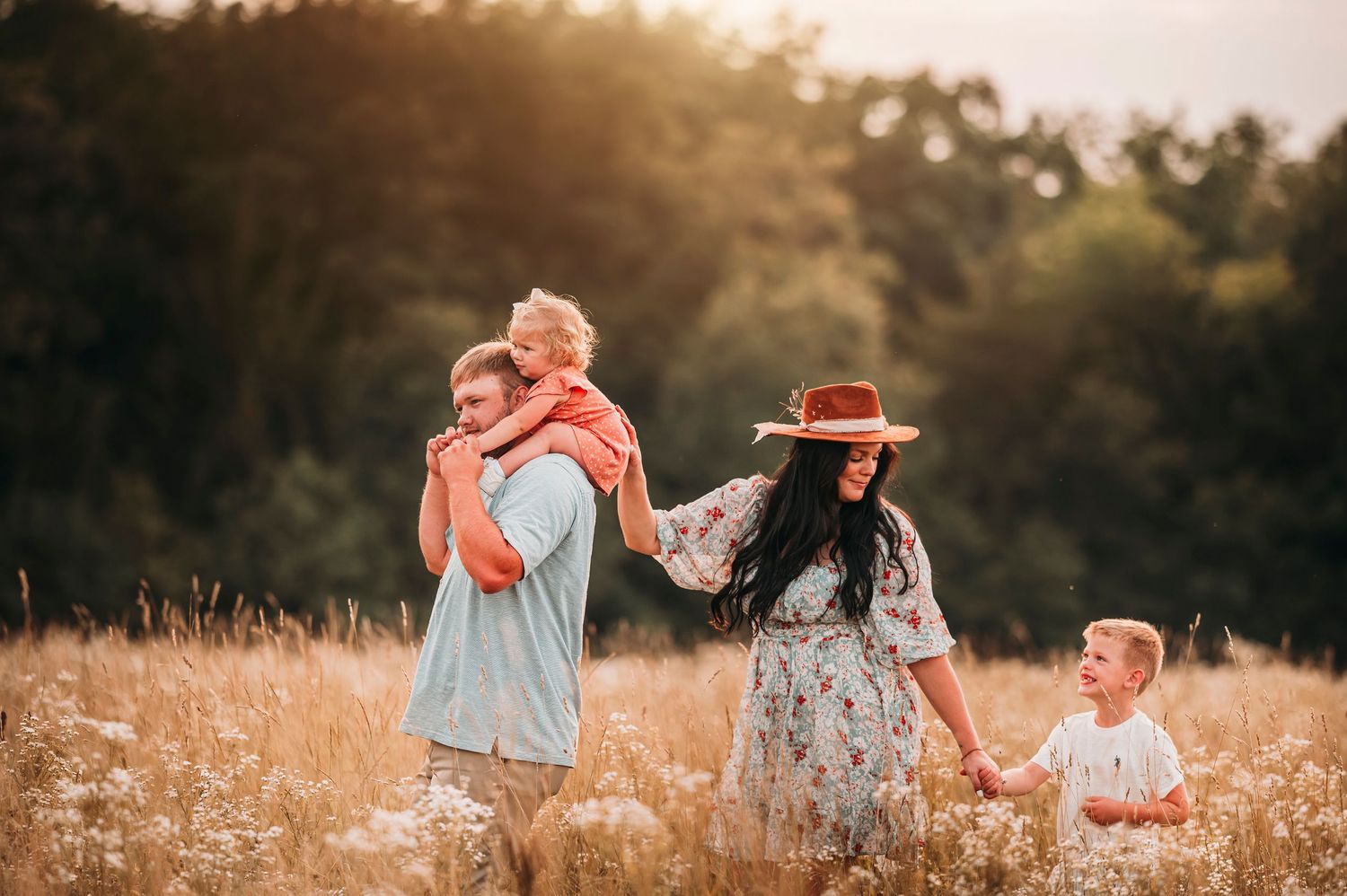 Happy family walking through a golden sunlit field, father carrying child on shoulders at sunset.