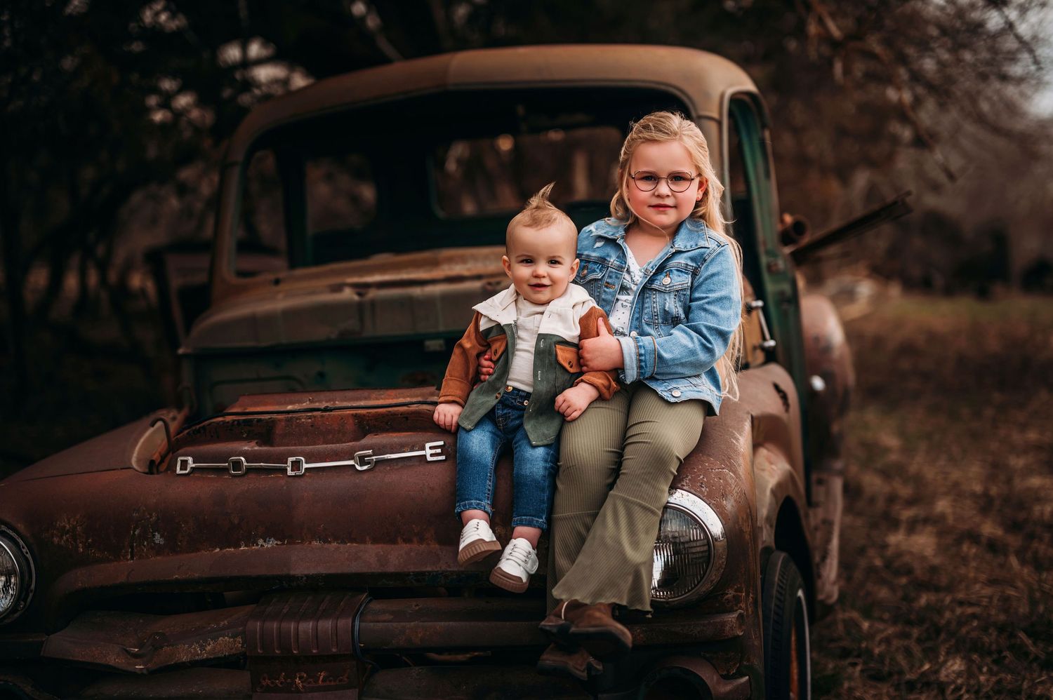 Two young sisters posing together on a rustic vintage truck in an outdoor autumn setting.