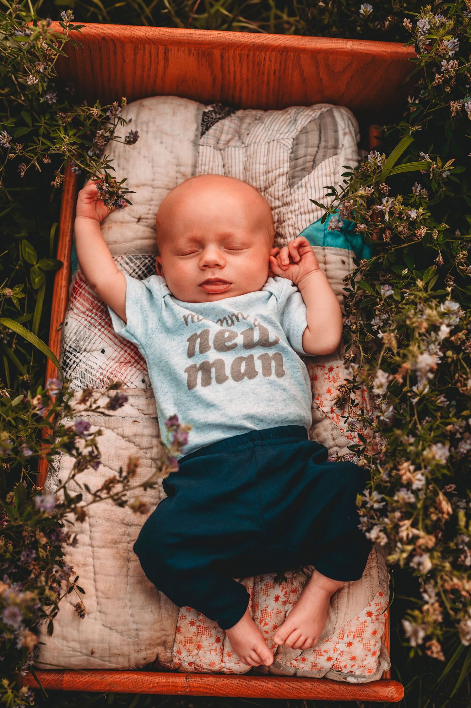 Newborn baby sleeping peacefully in a wooden crib surrounded by blooming wildflowers outdoors.