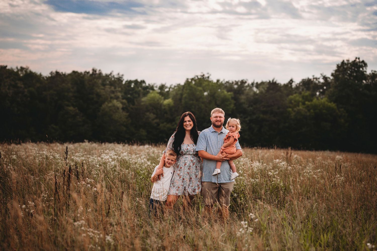 Smiling family of four posing together in a tall grass meadow under a cloudy sky.