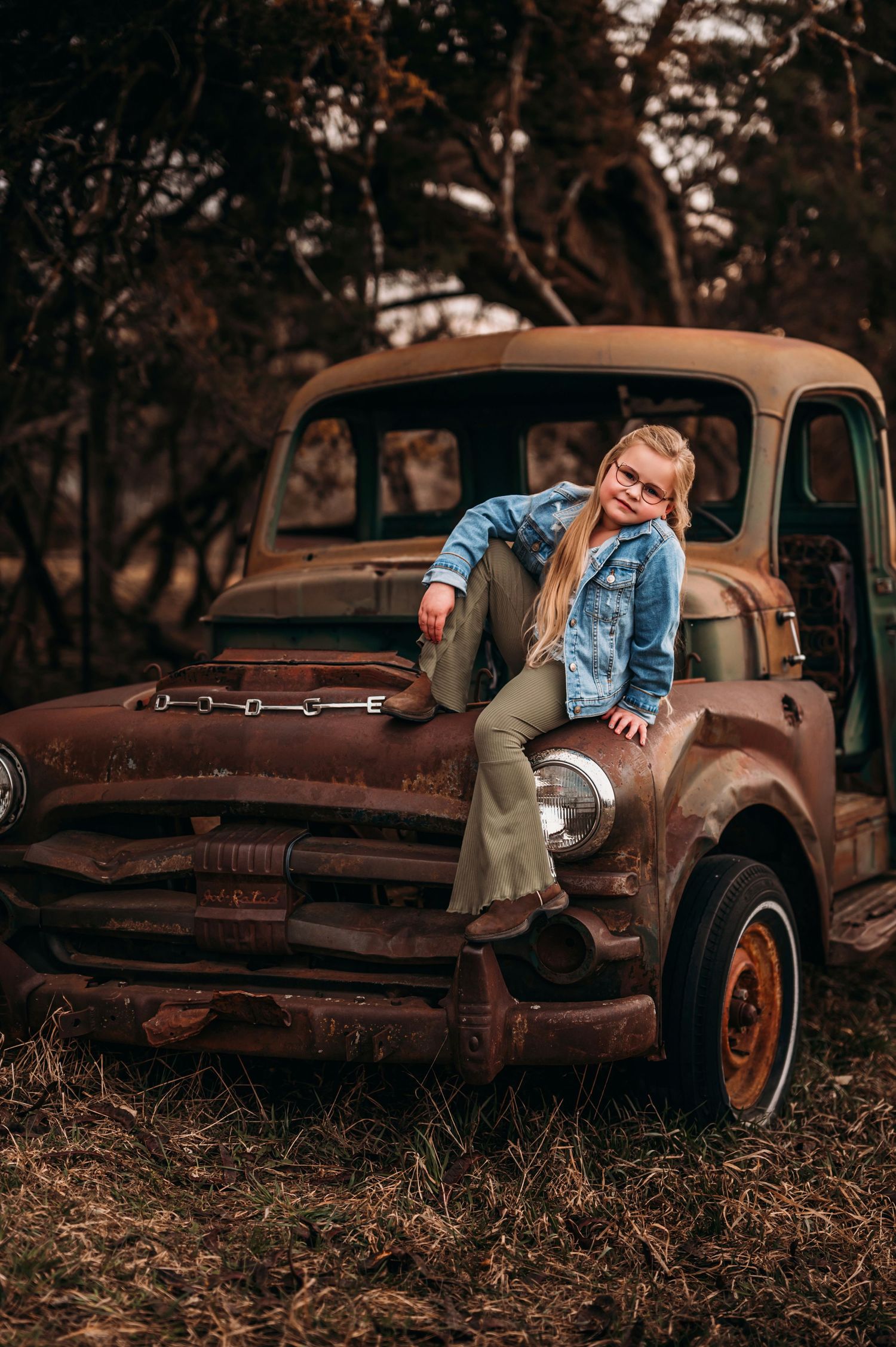Young blonde girl in denim jacket sitting on the hood of a rusty vintage truck in a field.