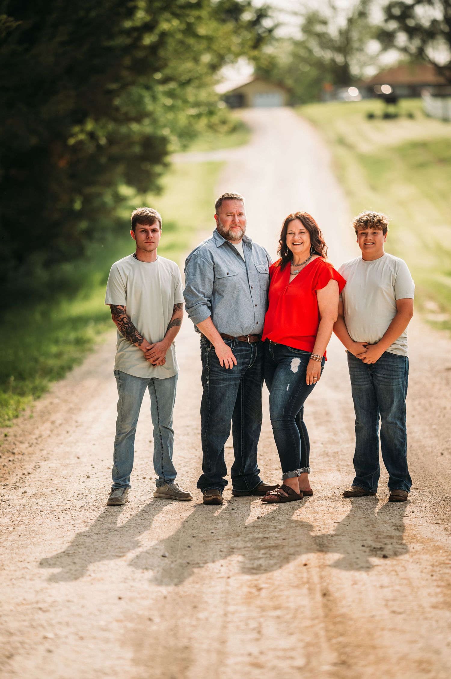 Family of four posing together on a rural dirt road surrounded by lush green summer trees.