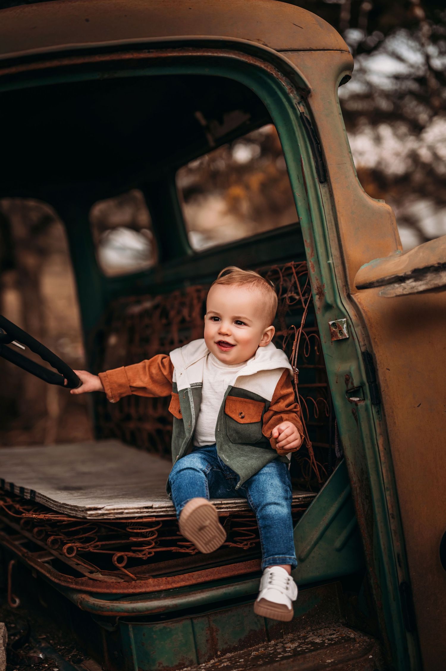 Happy toddler in brown jacket sitting in the open door of a vintage green truck outdoors.