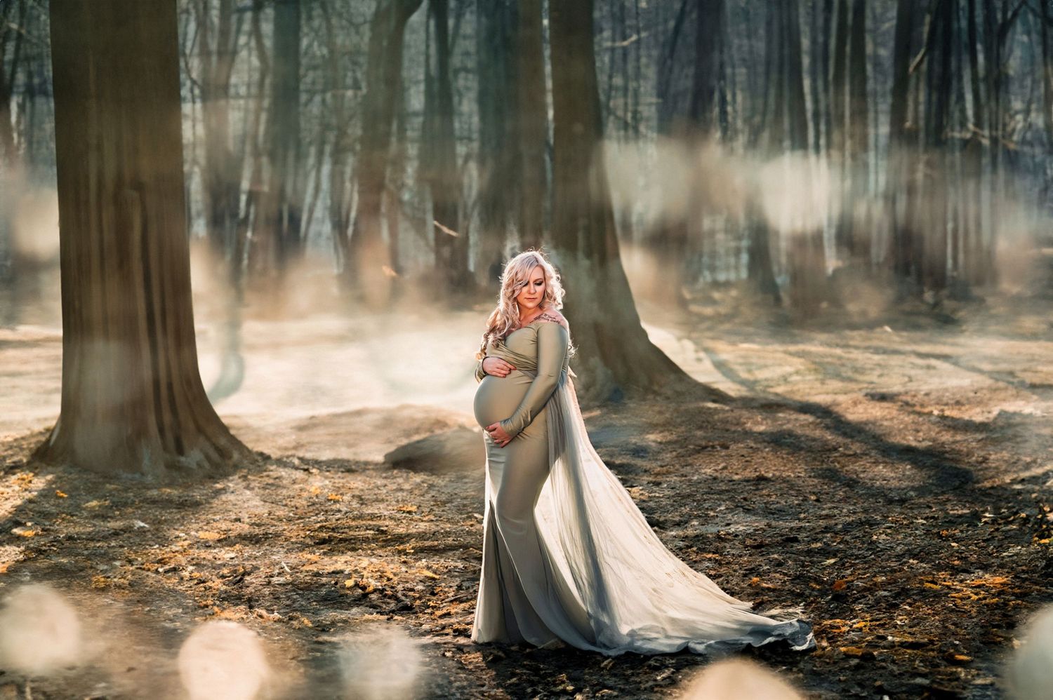 Pregnant woman in elegant silver gown standing in sunlit forest with dramatic light rays filtering through trees.